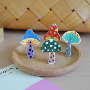 Peut inclure: Quatre écussons brodés en forme de champignons aux couleurs et motifs variés. Un est bleu avec une tige violette, un est rouge avec un dessous jaune, un est vert avec une tige beige, et un est bleu sarcelle avec une tige grise. Détails brodés.