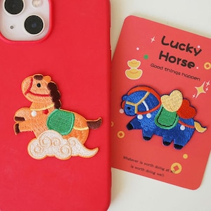 May include: A red phone case with an embroidered orange horse design and a red card with a blue horse design. The card reads "Lucky Horse. Good things happen" and "Whatever is worth doing at is worth doing well."