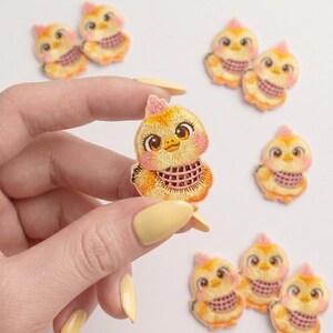 Puede incluir: Un pequeño parche bordado que muestra un pollito amarillo con grandes ojos marrones, una cresta rosa y una bufanda de cuadros rosas. El pollito tiene un cuerpo redondeado, pico naranja y mejillas sonrosadas. Varios parches idénticos están dispersos.
