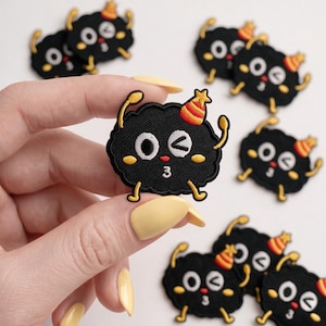May include: A black embroidered patch featuring a cartoon character with a round body, yellow arms and legs, and a red and yellow striped party hat. The character has one open eye, one winking eye, and a small red mouth shaped like the number 3.