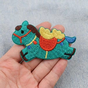 Op de afbeelding: Geborduurde patch van een turquoise carrouselpaard met een rood zadel en gele deken. Het paard heeft bruine en blauwe accenten en een rode teugel. Deze patch is geschikt voor het versieren van kleding of accessoires.