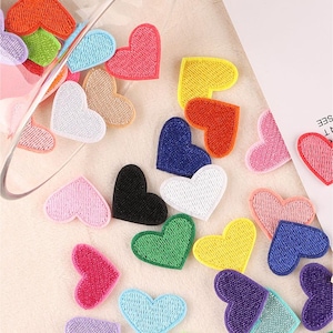 May include: A collection of colorful heart-shaped fabric patches in various colors, including red, pink, yellow, green, blue, and black. The patches are embroidered with a textured stitch and are scattered across a light-colored surface.