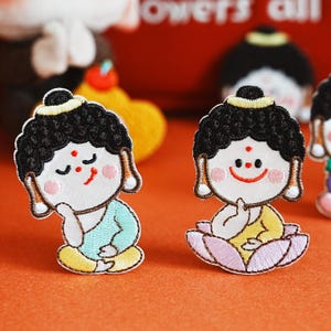 May include: Embroidered patches with a cartoon Buddha design. The patches have black hair, rosy cheeks, and are in a seated position. One patch is on a lotus flower, and the other is in a meditative pose. The patches have a yellow, blue, and pink colour scheme.