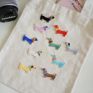 Puede incluir: Un bolso de tela beige con diez parches bordados de perros salchicha en varios colores, incluyendo negro, rosa, rojo, azul, verde, blanco, azul claro, amarillo y marrón, cada uno con un pequeño pañuelo de color alrededor del cuello.