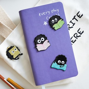 Puede incluir: Un cuaderno morado con el texto escrito a mano "every day counts" y una estrella. El cuaderno está decorado con personajes negros bordados leyendo libros. Una bolsa de mano blanca con el texto "FAVORITE HERE" está en el fondo.