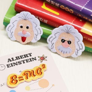 May include: Embroidered patches featuring Albert Einstein's likeness, one with his tongue out and the other wearing sunglasses. Also included is a patch with the formula E=mc². The patches are on a light pink card with the name "ALBERT EINSTEIN" printed on it.