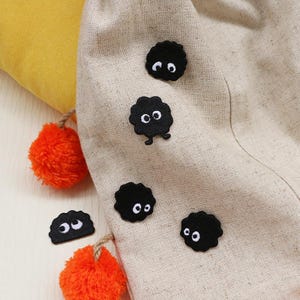 May include: A beige fabric bag decorated with several black, cartoon characters with white eyes. The bag is adorned with orange pom-poms and a yellow pillow. The characters have a fluffy, cloud-like appearance.