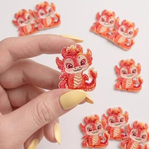May include: A small, embroidered patch featuring a cartoon dragon with red and orange scales, large eyes, and a light-colored belly. The dragon has small wings and a curled tail, with several identical patches arranged around it on a plain surface.