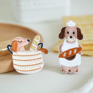 May include: Embroidered patches featuring cartoon dogs. One patch shows a dog in a cup with a baguette, the other a dog wearing a chef's hat and apron holding a loaf of bread. The patches are beige, brown, and white.