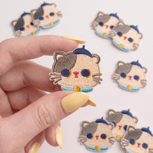 May include: Multiple embroidered cat patches featuring a beige face with grey patches, dark blue eyes, a small red nose, and a blue beret. Each cat wears a light blue collar with a yellow circular charm. The patches are small and detailed.