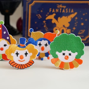 May include: Three embroidered clown patches featuring colourful designs. One patch has a green afro and orange collar, another has yellow hair with a blue hat, and the third has a purple hat. Each clown face displays black star-shaped eyes.