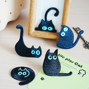 Peut inclure: Plusieurs écussons brodés noirs en forme de chat avec des yeux bleu vif. L'un des écussons est à l'intérieur d'un cadre en bois. Une carte verte avec le texte "life plus Cat" est également présente.