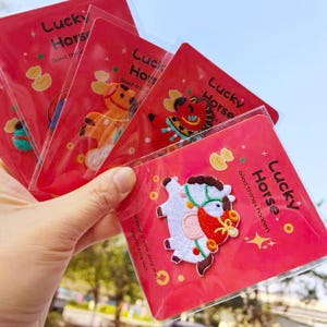 May include: A hand holding several red cards with embroidered horse designs. Each card has the text "Lucky Horse" and "Good things happen." The horse designs are in various colours, including white, orange, and red.