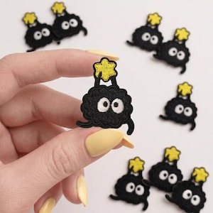 May include: A collection of small, black, circular embroidered patches featuring two large white eyes and a yellow star positioned above the head. Each patch has a textured, fuzzy appearance with two small limbs extending from the bottom sides.