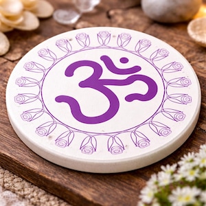 May include: A circular white coaster featuring a purple Om symbol and a floral border. The coaster is displayed on a wooden surface. Other decorative items are visible in the background.