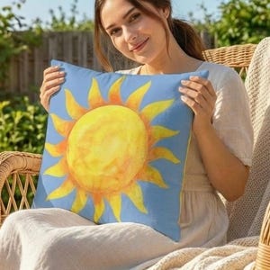 Puede incluir: Un cojín cuadrado azul con un diseño de sol en acuarela. El sol es amarillo con rayos naranjas. La persona está sentada en una silla de mimbre en una terraza de madera y sostiene el cojín. Hay una maceta con flores coloridas cerca.