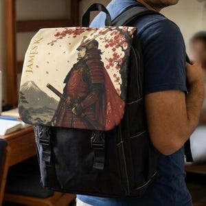 Könnte beinhalten: Ein schwarzer Rucksack mit einer Klappe, die eine Illustration eines Samurai in roter Rüstung unter einem Kirschblütenbaum zeigt, mit dem Berg Fuji im Hintergrund und dem Text JAMES K. vertikal auf der linken Seite der Klappe gedruckt.