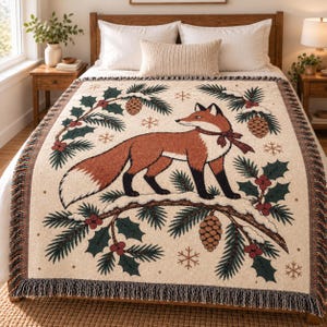 Könnte beinhalten: Eine gewebte Wohndecke mit einem zentralen orange-weißen Fuchs, der ein rotes Band um den Hals trägt. Das Design zeigt verschneite Tannenzweige, Tannenzapfen, Stechpalmenbeeren und Schneeflocken auf cremefarbenem Grund mit Fransen.