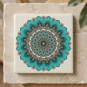 Puede incluir: Azulejo cerámico cuadrado con un diseño de mandala turquesa y coral. El mandala presenta un patrón detallado de puntos y círculos, creando una obra de arte simétrica e intrincada. El azulejo tiene un fondo color crema y se muestra sobre una superficie de piedra clara.