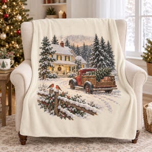 May include: A cream-coloured fleece throw blanket featuring a printed winter scene of a snow-covered house, a red vintage pickup truck carrying a Christmas tree, a wooden fence, and two small birds perched on a post in a snowy landscape.