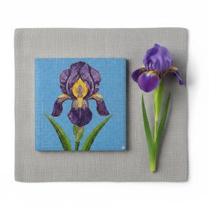 Puede incluir: Una impresión artística cuadrada con una ilustración detallada de una flor de iris púrpura con detalles amarillos y hojas verdes sobre un fondo de mosaico azul claro. Una flor de iris real está al lado de la impresión sobre una superficie gris.