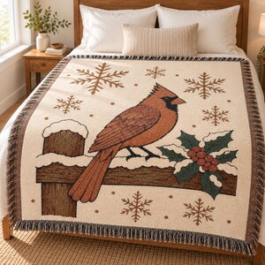Può includere: Una coperta tessuta con sfondo color crema che presenta un'immagine centrale di un cardinale rosso appollaiato su una staccionata in legno marrone. Il design include foglie di agrifoglio verdi con bacche rosse, fiocchi di neve marroni e frange.