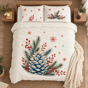 Könnte beinhalten: Ein cremefarbener Bettbezug mit zwei passenden Kissenbezügen und einem festlichen Weihnachtsmotiv. Das Design zeigt einen großen Tannenzapfen in der Mitte, umgeben von grünen Tannennadeln, roten Beeren sowie verstreuten roten Schneeflocken und goldenen Sternen.