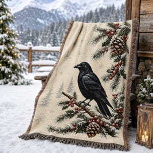 Puede incluir: Una manta tejida que presenta un cuervo negro posado en una rama de pino con bayas rojas y piñas sobre un fondo crema. El diseño incluye patrones sutiles de copos de nieve y un borde con flecos, mostrando una detallada escena invernal.