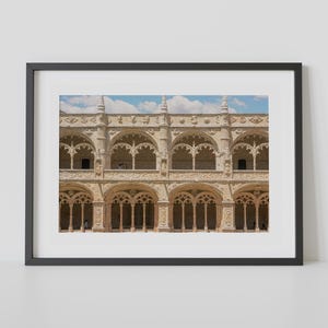 May include: Framed print of a detailed, ornate stone building facade. The building features arched windows and decorative carvings, with a glimpse of blue sky and clouds visible. The architecture suggests a historical or religious structure.