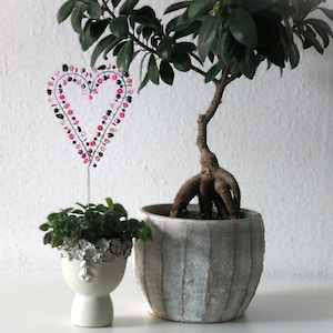 Könnte beinhalten: Ein kleiner Bonsai-Baum in einem strukturierten grauen Topf, neben einem kleinen weißen Pflanzgefäß mit Gesichtsdesign. Eine Draht-Herz-Dekoration mit rosa und schwarzen Perlen befindet sich in dem kleinen Pflanzgefäß.
