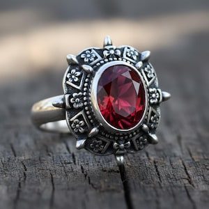 Könnte beinhalten: Ein silberner Ring mit einem ovalen, tiefroten Edelstein. Die Fassung des Rings ist aufwendig gestaltet und weist florale und geometrische Muster auf. Der Edelstein ist der Mittelpunkt, eingebettet in einen detaillierten, dekorativen Rahmen. Der Ring wird auf einer Holzoberfläche präsentiert.