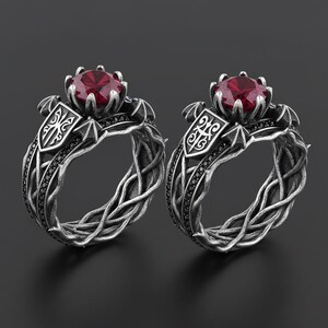 May include: Two silver rings with a gothic design. Each ring features a large, round, red gemstone set in a crown-like setting. The bands have a woven pattern and are accented with black stones. The rings have a shield-like emblem.