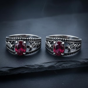 May include: Two silver rings with oval, deep red gemstones. The bands feature a bat and skull design with black accents. The rings are displayed on a dark, textured surface, creating a gothic or alternative aesthetic.