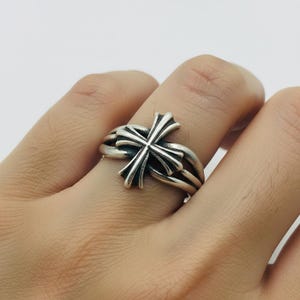 Könnte beinhalten: Ein silberner Ring mit einem detaillierten Kreuzdesign. Der Ring hat ein mehrschichtiges Band und ein markantes Kreuz-Mittelstück mit aufwendigen Details. Der Ring wird an einem Finger gezeigt.