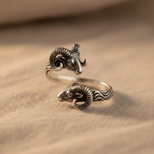May include: An open-style silver ring featuring two detailed ram heads facing each other. The metal has an oxidised finish to highlight the texture of the horns and facial features, with a wavy pattern engraved along the band of the ring.