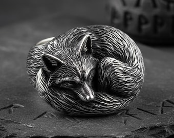 Puede incluir: Anillo plateado con un diseño detallado de un zorro enrollado. El zorro, con los ojos cerrados, descansa sobre una superficie oscura y texturizada. El anillo tiene un aspecto detallado y texturizado.