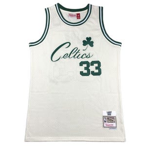 Può includere: Maglia da basket color crema con finiture e scritte verdi. La maglia presenta la scritta "Celtics" in verde, un trifoglio verde e il numero "33". È visibile un'etichetta "Hardwood Classics".