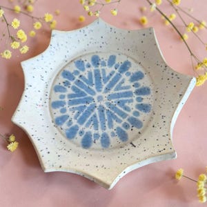 Puede incluir: Un plato de cerámica con forma de estrella y acabado moteado blanquecino. El centro presenta un diseño floral azul. El plato es una pieza decorativa para servir alimentos o exhibir objetos.