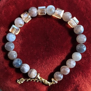 Peut inclure: Un bracelet de perles composé de perles rondes grises et de perles carrées transparentes. Le bracelet est doté d'un fermoir doré et est présenté sur une surface rouge. Les perles ont un aspect subtil et irisé.
