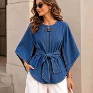 May include: A blue denim cape-style top featuring a round neckline with a decorative buckle closure, wide flared sleeves, and a matching fabric tie belt at the waist. The garment has a structured design with visible stitching and a curved hemline.