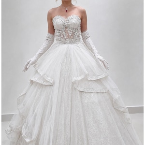 May include: A strapless white wedding gown with a sparkling bodice and layered skirt. The dress features intricate beadwork and is paired with long, white gloves. A tiara and necklace with a red gem complete the bridal look.