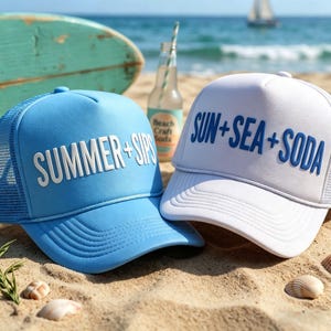 Può includere: Due cappellini da camionista su una spiaggia sabbiosa. Uno è azzurro con "SUMMER + SIPS" in bianco, l'altro è bianco con "SUN + SEA + SODA" in blu. Una bottiglia di soda e una tavola da surf sono sullo sfondo.
