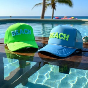 Puede incluir: Dos gorras de camionero con la palabra "BEACH" bordada en la parte delantera. Una gorra es de color verde brillante con la parte trasera de malla a juego, y la otra es azul claro con un detalle de cuerda blanca y la parte trasera de malla azul. Las gorras están sobre una superficie de madera cerca de una piscina.