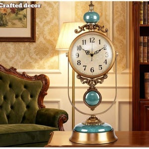 May include: An antique-style table clock with a gold-colored frame and a cream-colored clock face. The clock features teal accents and a decorative pendulum. The clock is displayed on a wooden table, with a green sofa and a bookshelf in the background.