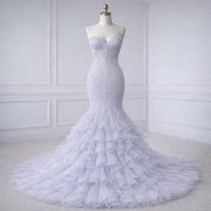 Puede incluir: Un vestido de novia blanco de sirena sin tirantes sobre un maniquí. El corpiño está detallado con encaje y abalorios. La falda presenta capas de tela con volantes que se extienden en una larga cola. El vestido se exhibe en una habitación blanca y luminosa.