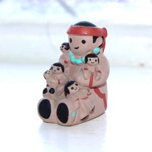 May include: A ceramic figurine depicting a Native American figure holding several smaller figures. The figurine has a red headband, turquoise necklace, and black hair. The base is black with rounded edges. The overall color palette is earthy tones.