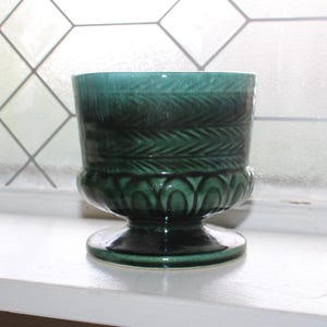 Peut inclure: Un cache-pot en céramique vert sarcelle foncé avec une finition brillante, présentant une section supérieure cylindrique ornée d'un motif de feuilles en forme de fougère et une base arrondie avec des détails cannelés sur un pied.