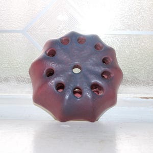 May include: A circular ceramic flower frog with a scalloped edge and a deep red and dark purple gradient glaze. The object features a central hole surrounded by a ring of ten smaller circular openings, designed for arranging stems in a vase.