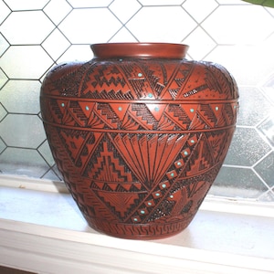 Może przedstawiać: Wazon ceramiczny w kolorze terakoty z misternymi, czarnymi, wytrawianymi wzorami geometrycznymi i małymi, inkrustowanymi kropkami w kolorze turkusowym. Naczynie ma szeroki, zaokrąglony korpus z krótką, wąską szyjką i gładką, płaską krawędzią.