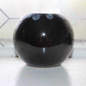 May include: A round, black ceramic vase with a glossy finish. The vase has a wide opening and a slightly textured rim with white accents. It sits on a white surface, reflecting the light. The vase is a decorative item.